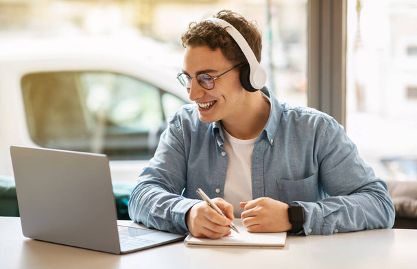 Mosolygó fiatal európai fickó szemüvegben, fejhallgató laptoppal, jegyzetel, dolgozik, tanul vagy online kommunikáció kávézóban. Online találkozó, kommunikáció, lecke és webinárium - Fotó, kép