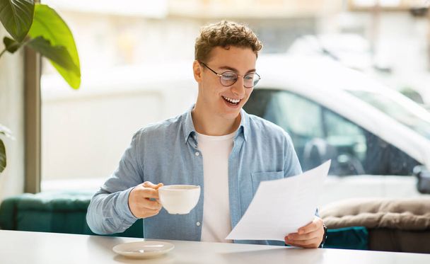 Hymyilevä tuhatvuotinen eurooppalainen mies opiskelija johtaja työtä, opiskella, lukea asiakirja, juoda kahvia modernissa kahvilassa, panoraama. Paperityöt, elämäntapa ja aamiainen, startup-yrityksen suunnittelu - Valokuva, kuva
