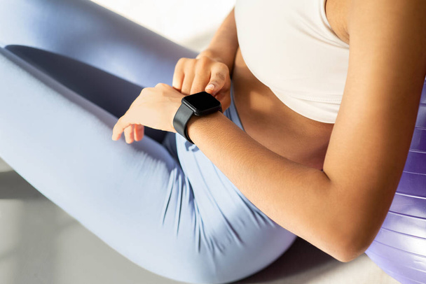 Крупный план фитнес-ориентированной женщины, проверяющей свои умные часы после тренировки, сидящей на мяче для упражнений, символизирующий технически продуманный подход к отслеживанию здоровья, обрезанной - Фото, изображение