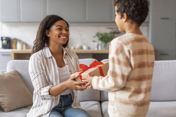 Küçük siyah çocuk, mutlu annesine kırmızı kurdeleli hediye kutusu veriyor, doğum günüyle ya da anneler günüyle karşılıyor, iki Afro-Amerikan ailesini seven, evlerinde samimi bir değiş tokuş gerçekleştiren - Fotoğraf, Görsel