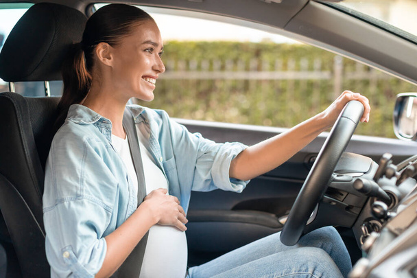 Szczęśliwa ciężarna kobieta prowadzi nowoczesny samochód siedząc z jedną ręką na brzuchu, a drugą na kierownicy wewnątrz samochodu. Kobieta w ciąży poruszająca się pojazdem, zapewniająca bezpieczeństwo i komfort podczas jazdy - Zdjęcie, obraz