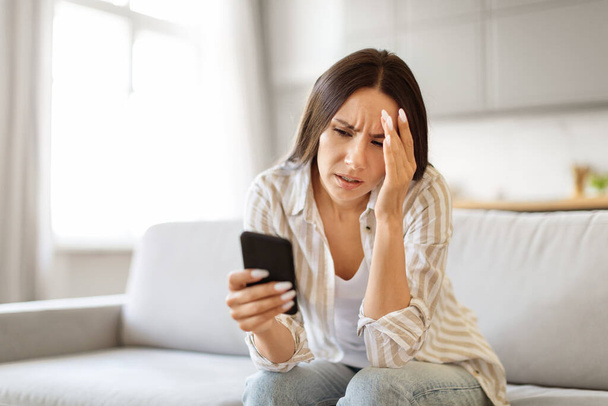 Zaniepokojona młoda kobieta patrząca na ekran smartfona i dotykająca głowy, siedząca na kanapie w domu, sfrustrowana Europejka czytająca nieprzyjemne smsy, emocjonalnie reagująca na złe wiadomości, wolna przestrzeń - Zdjęcie, obraz