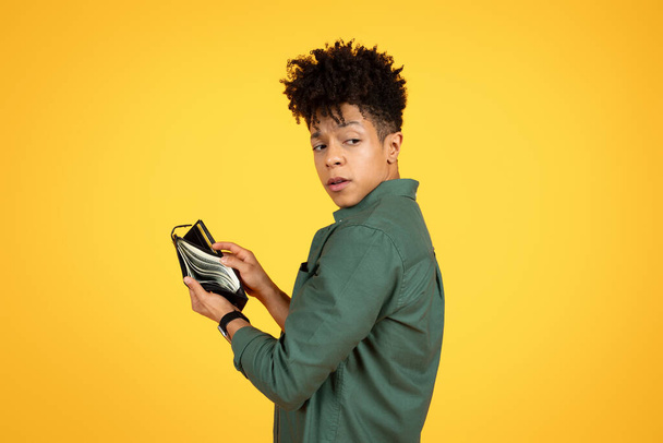 Theft Concept. Suspicious african american young man stole somebody wallet and counting his loot. Yellow background with copy space, Greedy black guy decides to buy something - Photo, Image