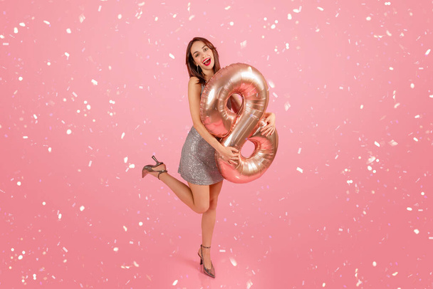Femme joyeuse dans une robe pailletée tenant un ballon numéro 9 en or rose avec des confettis tombant sur un fond rose, célébrant une occasion festive. Fête du printemps - Photo, image