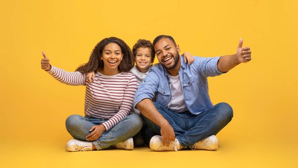 Как Концепция. Счастливая афроамериканская семья из трех человек, показанных на камеру, сидя на жёлтом фоне в студии, счастливые чёрные родители и маленький сын одобряют или рекомендуют, скопировать пространство - Фото, изображение