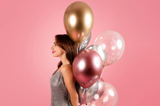 Una mujer serena con un vestido plateado brillante mira a lo lejos, su perfil acentuado por un montón de elegantes globos metálicos sobre un cálido fondo rosa. Evento de celebración - Foto, Imagen