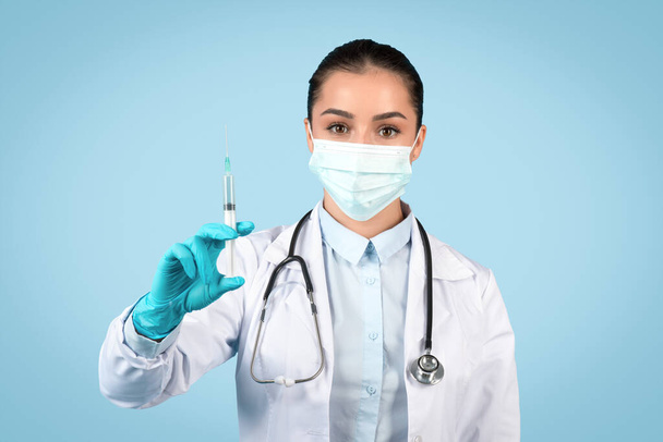 Poważny personel medyczny kobiety w masce chirurgicznej i rękawiczkach ostrożnie wyświetla strzykawkę, gotową do szczepienia lub leczenia, niebieskie tło - Zdjęcie, obraz