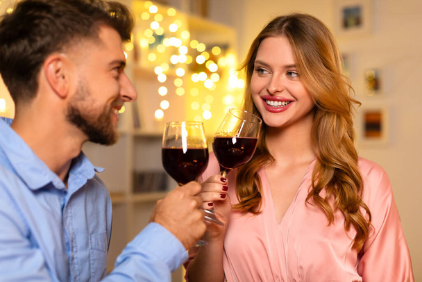 Giovane coppia sorridente in brindisi allegro con bicchieri di vino rosso, condividendo momento di felicità in uno sfondo di luci fiabesche calde e festive - Foto, immagini