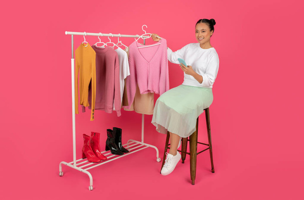 Personal Stylist App. Happy Asian Woman With Smartphone Sitting On Chair Near Clothing Rack, Smiling Korean Female Fashion Consultant Demonstrating New Clothes, Posing On Pink Background - Photo, Image