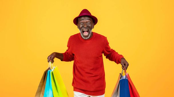 Shocked excited old black man shopaholic in hat with open mouth and many bags scream, isolated on orange studio background, panorama. Shopping wow, huge sale at store, emotions - Photo, Image