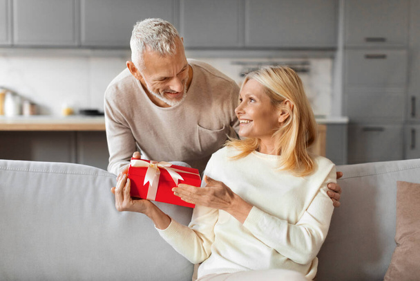 Der grauhaarige Mann begrüßt seine schöne lächelnde Frau mit einem Geschenk zum Valentinstag. Glücklich verheiratete Senioren feiern Jubiläum, Weihnachten und tauschen zu Hause Geschenke aus - Foto, Bild