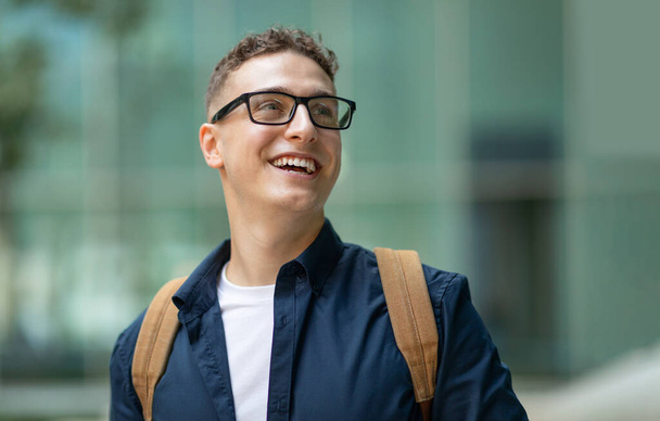Cheerful european student with glasses and a backpack looking up and smiling, standing in front of a modern glass building, wearing a dark jacket over a white shirt, close up - Photo, Image