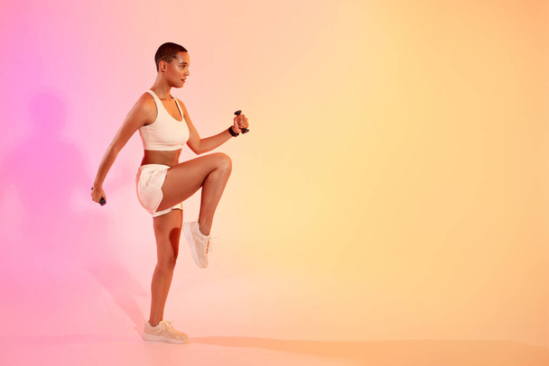 Grave giovane donna dinamica in marcia atletica esegue un esercizio ad alto ginocchio con un'espressione concentrata su uno sfondo morbido rosa e giallo ombre. Vestibilità, sport e assistenza sanitaria - Foto, immagini