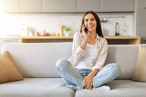 Happy young woman chatting on cellphone, sitting cross-legged on sofa at home, cheerful beautiful female enjoying mobile phone conversation while relaxing in living room, copy space - Photo, Image