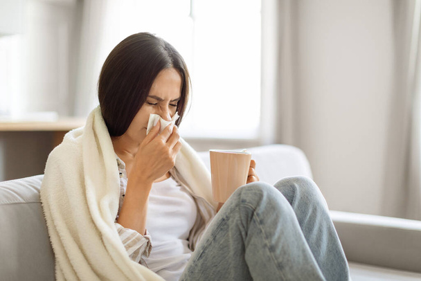 Mulher jovem sentindo-se doente em casa, soprando o nariz com um tecido e segurando caneca com chá, fêmea millennial doente envolto em cobertor sentado no sofá na sala de estar interior, sofrendo gripe sazonal - Foto, Imagem