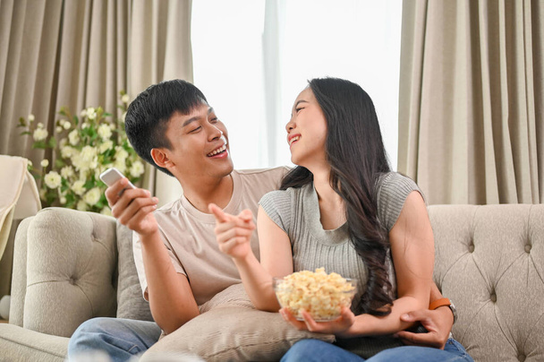 Onnellinen nuori aasialainen pari syö popcornia nauttien television katselusta olohuoneen sohvalla yhdessä.. - Valokuva, kuva