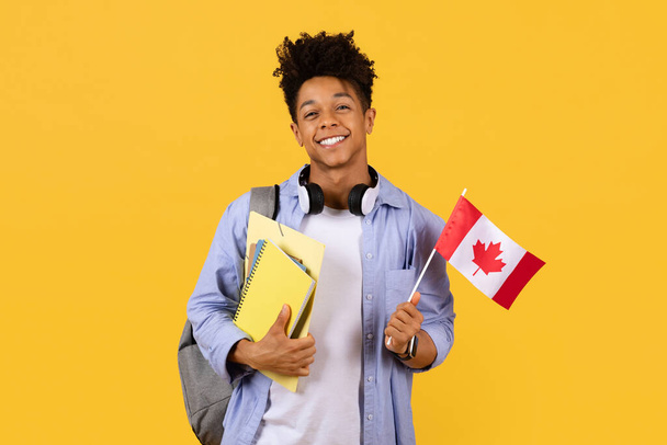 Allegro studente maschio nero che tiene orgogliosamente la bandiera canadese, indossa le cuffie, pronto per lo studio all'estero, sullo sfondo giallo, simboleggia la diversità e l'apprendimento delle lingue straniere - Foto, immagini