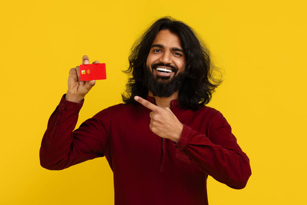 Joyeux excité beau barbu jeune homme indien hipster pointant vers la carte de crédit en plastique rouge dans sa main, recommandant sa banque, isolé sur fond jaune. Facilité bancaire virtuelle, paiement sans contact - Photo, image