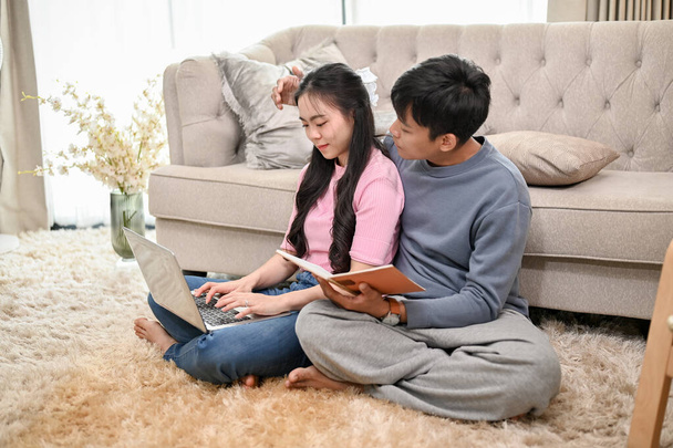 Een lieve en zorgzame jonge Aziatische man helpt zijn vriendin met haar werk, werkt thuis, zit samen op een tapijt in de woonkamer. - Foto, afbeelding