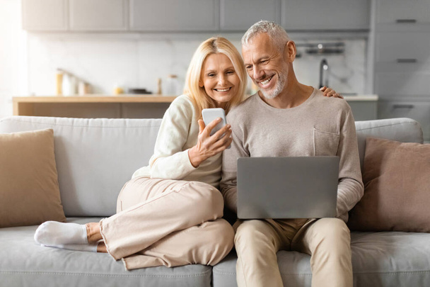 Rentner europäisches Paar, das Zeit zu Hause verbringt, mit Gadgets, Glückliche ältere Männer und Ehefrauen, die auf der Couch sitzen, Laptop und Smartphone benutzen, Fotos checken, Websurfen, Kopierraum - Foto, Bild