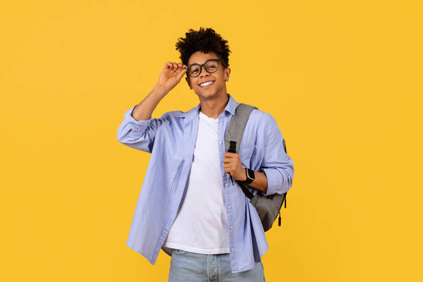 Smart aussehender junger schwarzer Student mit lockigem Haar justiert seine Brille und trägt dabei einen Rucksack, der vor leuchtend gelbem Hintergrund Zuversicht ausstrahlt. - Foto, Bild