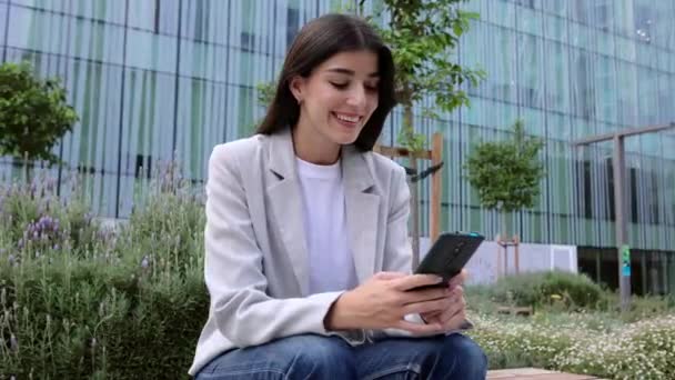 Επιτυχημένη νεαρή ενήλικη επιχειρηματίας που χρησιμοποιεί το κινητό τηλέφωνο χαλαρώνοντας πάνω από το κτίριο των επιχειρήσεων. Τρόπος ζωής της τεχνολογίας και επιχειρηματική ιδέα - Πλάνα, βίντεο