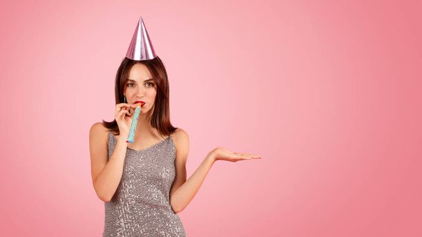 Playful woman in a sparkling dress and party hat blowing a party horn, with a hand extended as if to show something, against a festive pink background. Holiday celebration event - Photo, Image