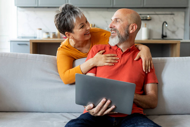 Ευτυχισμένο ζευγάρι ηλικιωμένων με φορητό υπολογιστή που διασκεδάζουν μαζί στο σπίτι, χαρούμενα ηλικιωμένοι σύζυγοι που χρησιμοποιούν υπολογιστή ενώ χαλαρώνουν στον καναπέ στο σαλόνι, γυναίκα αγκαλιάζει τον σύζυγο από πίσω, ελεύθερος χώρος - Φωτογραφία, εικόνα