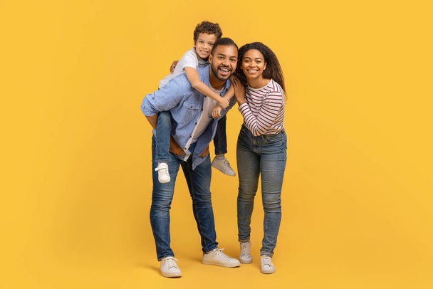 Full length shot of happy black family of three with preteen son having fun together over yellow background in studio, smiling african american parents and male kid sharing moment of joy, copy space - Photo, Image