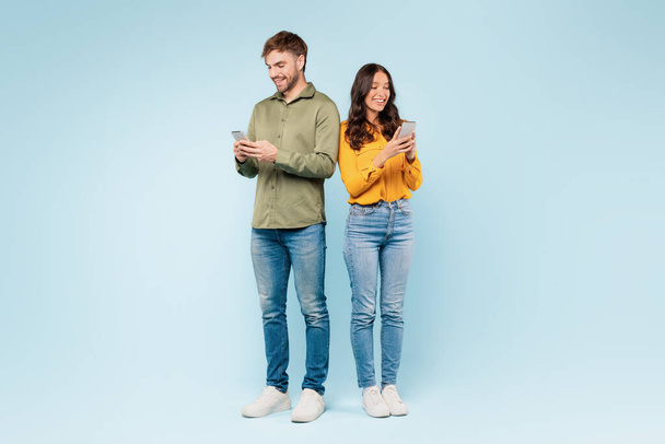 Tevreden echtpaar elk gericht op hun smartphones, sms 'en en communiceren met hun digitale wereld terwijl ze naast elkaar staan, tegen een blauwe achtergrond, volledige lengte - Foto, afbeelding