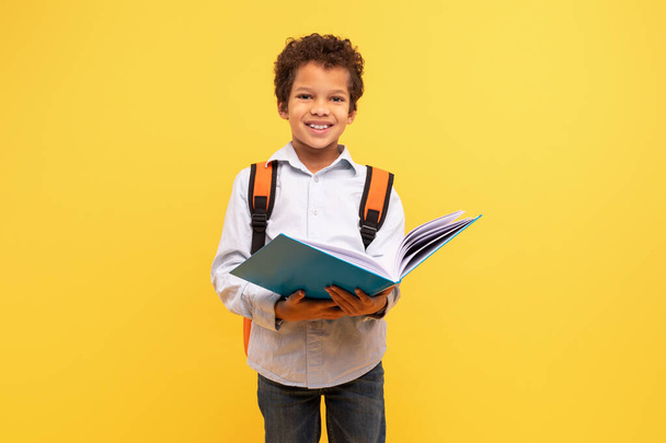 Intelligent black schoolboy engrossed in reading book, wearing an orange backpack smiling at camera against vibrant yellow background - Photo, image