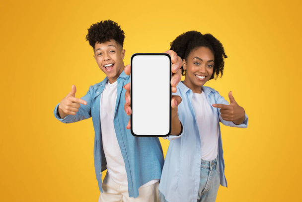 Щасливий впевнений молодий афроамериканський чоловік і жінка в випадкових точках пальців на смартфоні з порожнім екраном, ізольовані на помаранчевій фоновій студії. Веб-сайт, презентація додатків, рекомендації - Фото, зображення