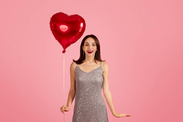 Surprised and delighted glad caucasian woman in a glamorous silver sequin dress holding a heart-shaped red balloon, with an open gesture on a pink backdrop, studio, ad and offer - Photo, Image