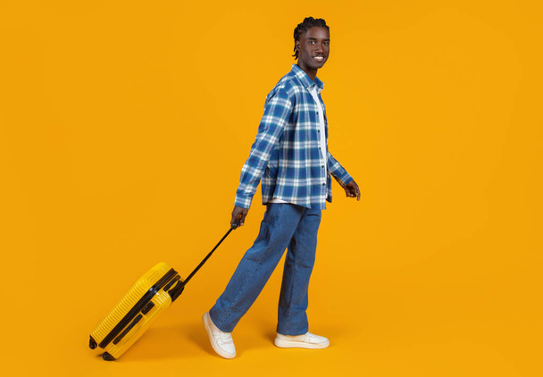 Uśmiechnięty młody czarny mężczyzna spacerujący z żółtą walizką, szczęśliwy facet ubrany w koszulę w kratę i dżinsy, gotowy na przygody, pozujący na jasnym pomarańczowym tle, cieszący się podróżami - Zdjęcie, obraz
