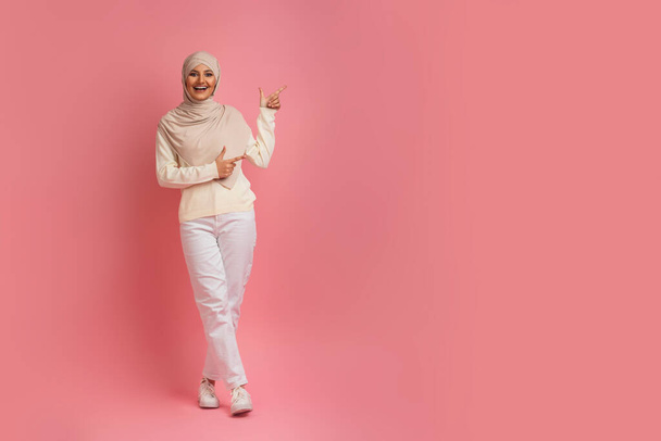 Donna allegra in Hijab che dimostra spazio di copia su sfondo rosa, sorridente giovane donna araba che indossa il velo a parte con le dita, mostrando il posto libero per la pubblicità, lunghezza completa - Foto, immagini