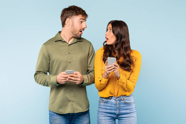 Εντυπωσιασμένος νεαρός άνδρας και γυναίκα κρατώντας ο καθένας smartphones, αντιδρώντας σε εκπληκτικά ή συναρπαστικά νέα, κοιτάζοντας ο ένας τον άλλο σε γαλάζιο φόντο - Φωτογραφία, εικόνα