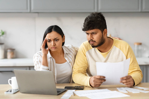 Mortgage problems concept. Overwhelmed young indian couple sitting together at kitchen, using laptop, holding papers documents, filling online form from home, have difficulties with bureaucracy - Photo, Image