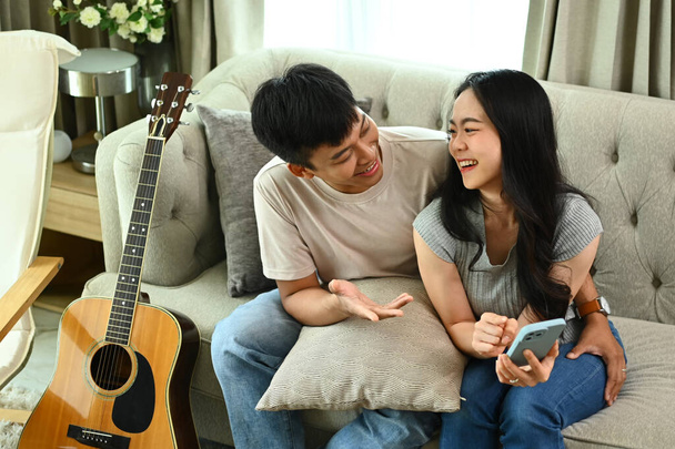 Fröhliches junges asiatisches Paar mit Smartphone auf bequemer Couch im Wohnzimmer. - Foto, Bild