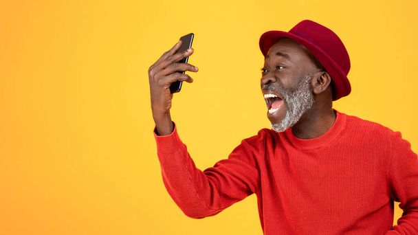 Щасливий або розлючений старший афроамериканський чоловік у капелюсі та червоному одязі, з відкритим ротом, кричить по телефону, ізольований на помаранчевому студійному фоні, крупним планом, панорама. Перемога, хороші новини, сварка - Фото, зображення