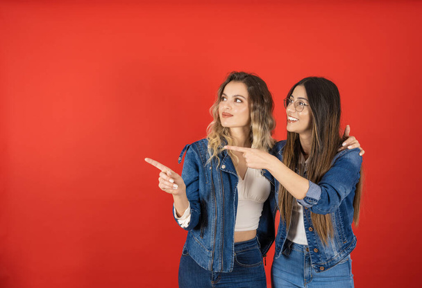 Point index finger aside, portrait of young female friends smiling point index finger aside. Showing copy space. Recommending concept idea. Standing together, wear denim jacket and jeans.  - Photo, Image