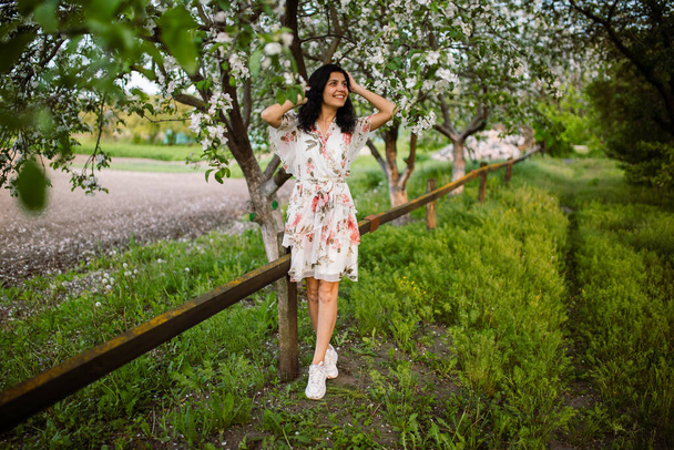Очаровательная счастливая девушка с длинными вьющимися волосами улыбается и смеется на фоне цветущих яблоневых вишневых деревьев в сельском парке весной - Фото, изображение