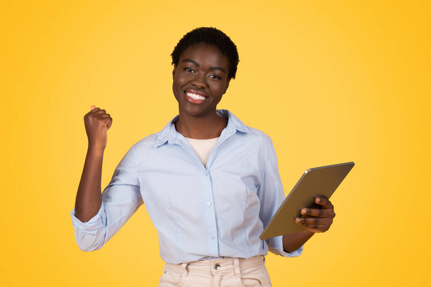 Blij jonge Afrikaanse Amerikaanse dame student in casual show tablet vieren overwinning, succes, genieten van online les, geïsoleerd op gele studio achtergrond. Studie en onderwijs, online app - Foto, afbeelding