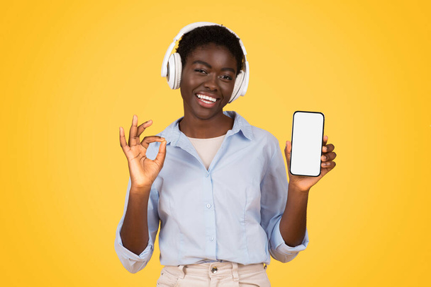 Glad young african american woman student in wireless headphones, listen music, show smartphone with empty screen, enjoy online lesson, isolated on yellow studio background. Study and education - Photo, Image