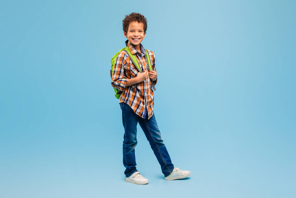 Happy schoolboy in plaid shirt and jeans with green backpack, standing confidently with smile against refreshing blue background - Photo, Image