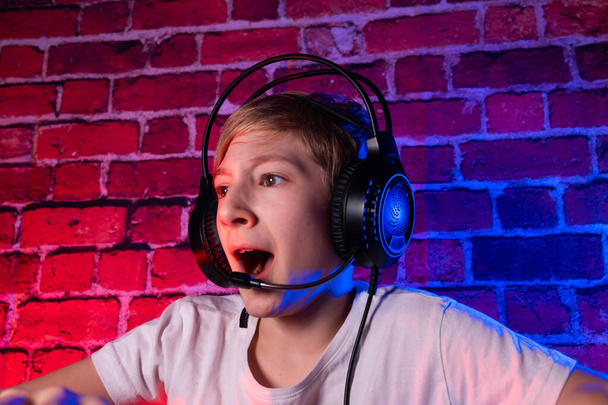 Ein fröhlicher Teenager an einer Tastatur mit Kopfhörern, der am Schreibtisch Videospiele spielt. Ein Gamer-Junge spielt Computerspiele gegen eine neonbeleuchtete Ziegelwand. - Foto, Bild