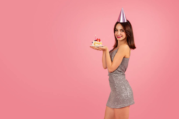 Сияющая женщина с каштановыми волосами в блестящем серебряном платье для вечеринки и в конической шляпе держит праздничный торт с зажженной свечой на розовом фоне. Праздничное мероприятие - Фото, изображение