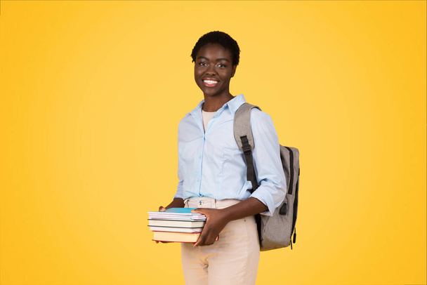 Positive young african american lady student with books, backpack, enjoy school lifestyle, isolated on yellow background, studio. Education, study, ad and offer, prepare to exam - Photo, Image