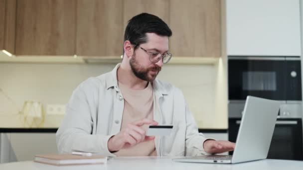 Homme caucasien concentré réécriture numéro de carte dans un ordinateur portable moderne tout en étant assis sur le lieu de travail personnel à la maison. Homme souriant vêtu de vêtements décontractés et de lunettes travaillant à l'intérieur. - Séquence, vidéo