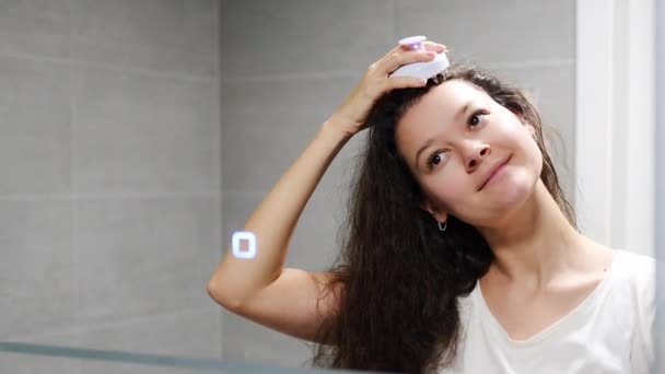 Young woman with dark curly hair doing self hair scalp massage with scalp massager or hair brush for hair growth stimulating at home bathroom. Reflected view of the mirror. High quality photo - Footage, Video
