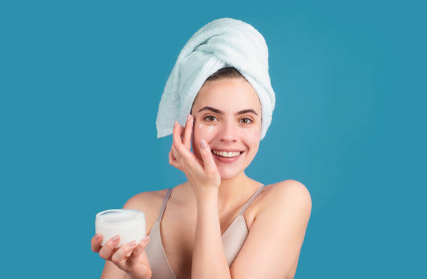 Beautiful happy girl with clean fresh skin. Facial treatment. Cosmetology beauty and spa. Girl apply skincare cream on face, woman wrap towel on head put facial creme on doing morning healthy skin - 写真・画像